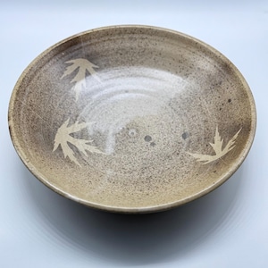 May include: A round ceramic bowl with a speckled, tan exterior and a lighter interior. The bowl features three leaf-shaped cutouts around the rim. Suitable for serving food or as a decorative piece.
