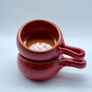 Puede incluir: Dos cuencos de cerámica roja apilados, con asas. Los cuencos tienen un acabado brillante y un interior marrón contrastante. Las asas son curvas y están integradas en el diseño del cuenco. Los cuencos son probablemente para servir sopa u otras porciones pequeñas.