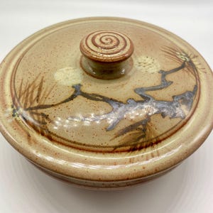May include: A ceramic lidded bowl with a tan and brown glaze. The lid has a spiral knob and is decorated with a branch and floral design in brown and grey. The bowl is suitable for serving or storage.