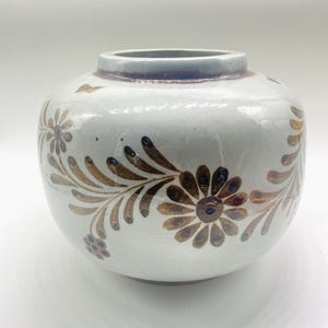 Peut inclure: Un vase rond en céramique blanc cassé avec une large ouverture et une surface légèrement texturée. Le vase est décoré de motifs floraux et d'accents bruns. Le bord est d'une nuance de brun plus foncée. C'est un objet décoratif.