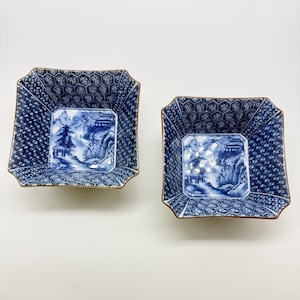 May include: Two square blue and white ceramic dishes with a landscape scene. The dishes have a geometric pattern around the edges and a brown rim. The landscape scene features mountains, trees, and buildings. The dishes are approximately 15 cm square.