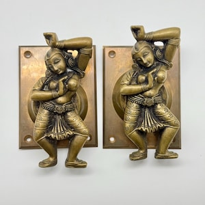 Puede incluir: Dos manijas de puerta antiguas de bronce con una figura detallada en una pose de baile. Las figuras están unidas a placas traseras rectangulares de bronce con un diseño circular. Las manijas tienen un color marrón dorado cálido y están sobre un fondo blanco.