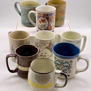 May include: A collection of ceramic mugs in various colors and designs. The mugs feature floral, landscape, and abstract patterns. Some mugs have a solid color interior, while others have patterned exteriors. The mugs are of different shapes and sizes.