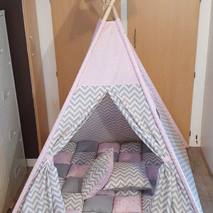 Könnte beinhalten: Ein rosa-graues Tipi-Zelt für Kinder mit Chevron-Muster und sternenbesetztem Stoff. Das Zelt hat einen gepolsterten Boden mit grauen und rosa Quadraten und wird mit zwei grauen und rosa Kissen geliefert.