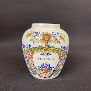 May include: A small, white ceramic vase with a colorful floral design and the word "VIRGINIA" in a central cartouche. The vase features a round body and a narrow opening, with a black background.