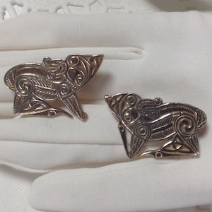 May include: Two silver-toned brooches in the shape of stylized animals with intricate details. The brooches are on a white surface.