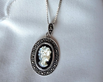 May include: Silver-toned necklace with an oval pendant featuring a cameo profile. The cameo is white against a black background, surrounded by small, dark stones. The necklace is displayed on a white surface.
