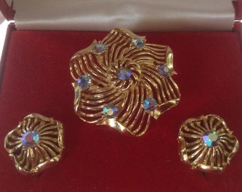 May include: A vintage gold-tone jewelry set featuring a brooch and matching clip-on earrings. The brooch and earrings have a floral design with iridescent, multi-colored rhinestones. The set is presented in a red box with a white interior.