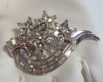 May include: A silver-toned brooch with a floral design. The brooch features clear, sparkling crystals arranged in a cluster of star-shaped flowers and rectangular accents. The brooch has a curved, elegant shape.