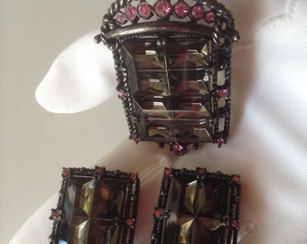 May include: A set of vintage jewelry featuring a large pendant and a pair of earrings. The pendant and earrings are made of metal with a dark finish and feature a large, faceted, clear glass stone surrounded by smaller pink stones.