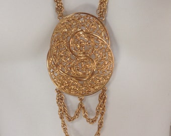 May include: A gold-tone necklace with a large, ornate pendant. The pendant is a filigree design with a swirling pattern. The necklace is made of multiple chains.