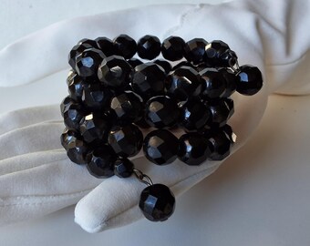 May include: A black beaded wrap bracelet with multiple strands of faceted beads. The bracelet is displayed on a white glove, showcasing the jewelry's dark color and design. The beads have a glossy finish, reflecting light.