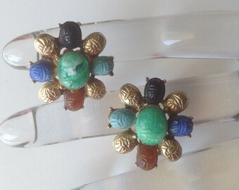 May include: A pair of gold tone earrings with a flower design. The earrings are made with green, blue, and brown stones. The stones are carved with scarab beetles.