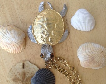 May include: A gold and silver turtle pendant necklace with a gold chain. The turtle is surrounded by seashells on a wooden surface.