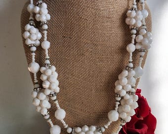 May include: A white beaded necklace with floral clusters and small silver accents, displayed on a brown jewelry bust. The necklace features multiple strands and a variety of bead sizes, creating a layered effect. A red rose is visible in the background.