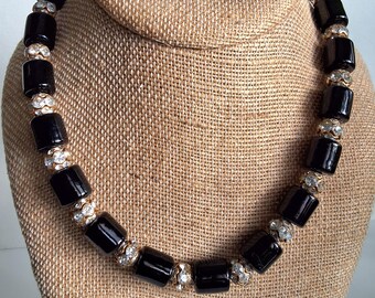 May include: A black beaded necklace with rectangular beads and rhinestone accents. The necklace is displayed on a tan jewelry bust. The beads are glossy and evenly spaced, with the rhinestone accents adding sparkle.