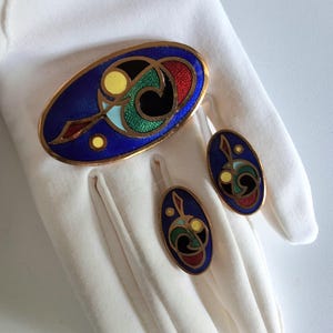 May include: A gold-toned brooch and a pair of matching oval earrings featuring cloisonné enamel work. The jewelry pieces display a deep blue background with abstract geometric patterns in yellow, green, red, black, and light blue enamel accents.