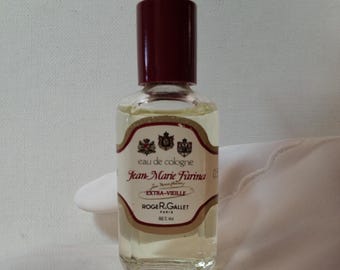 May include: A clear glass bottle of Jean-Marie Farina Eau de Cologne with a dark red cap. The label features ornate text and crests. The bottle contains a light yellow liquid and is held against a white background.
