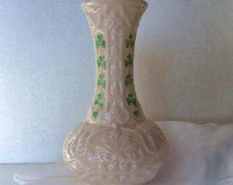 May include: A cream-colored ceramic vase with a bulbous base and a slender neck. The vase features embossed floral patterns and green shamrock designs. The vase is approximately 7 inches tall.