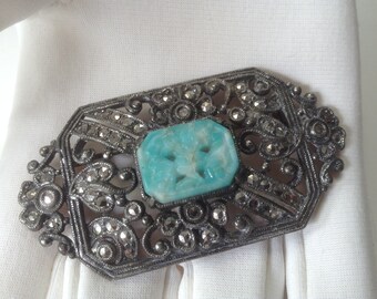 May include: Vintage silver-tone brooch with a rectangular turquoise-colored stone center. The brooch has an ornate filigree design with small, sparkling accents. The brooch is set against a white background.
