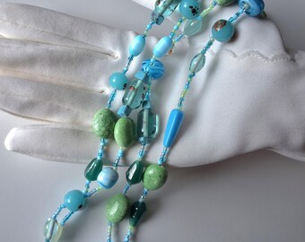 May include: A multi-strand necklace featuring a variety of blue and green beads. The beads are in various shapes, including round, oval, and oblong, and are strung on a delicate blue cord. The necklace is displayed on a white surface.