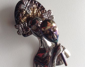 May include: A silver and gold toned brooch featuring a woman's face with a butterfly design on her head and a mask in her hand.