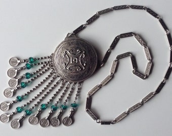 May include: A silver necklace with a round pendant featuring an intricate design. The pendant has a tassel of silver chains and green beads.