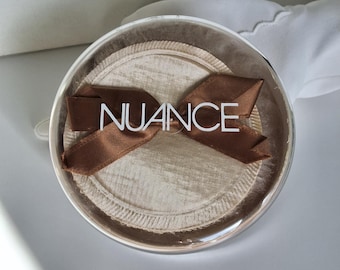 May include: A clear, round container with a brown ribbon and the word "NUANCE" in white. The container holds a beige, circular item with a textured surface. The ribbon is tied in a bow.
