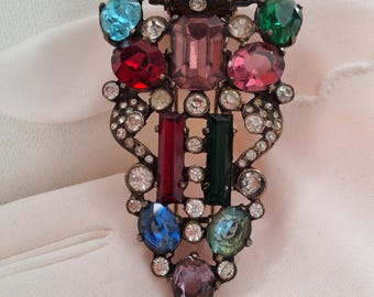 May include: A vintage brooch with an ornate design, featuring an array of colorful gemstones. The brooch showcases a rectangular blue stone at the top, surrounded by various shapes and colors, including red, pink, green, and blue. Small clear crystals add sparkle.