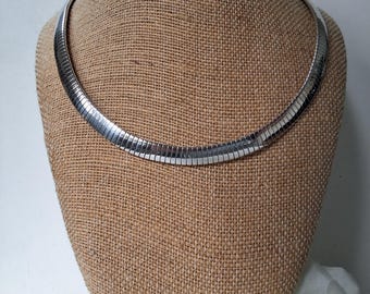 May include: A silver-colored necklace with a flat, ribbed design. The necklace is displayed on a tan mannequin head. The jewelry is a choker style, and the metal has a shiny finish.