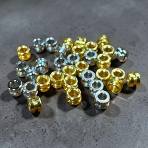 Op de afbeelding: Een verzameling kleine, cilindervormige metalen kralen in goud- en zilverkleurige tinten. Elke kraal heeft een gladde, ronde opening in het midden en een duidelijke horizontale groef die rond de buitenomtrek van het metalen lichaam loopt.