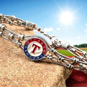 May include: Silver-toned bracelet featuring a Texas Rangers logo charm. The charm is round with a red, white, and blue design, displaying a "T" and the team name. The bracelet is made of multiple strands of metal and is displayed on a rock.