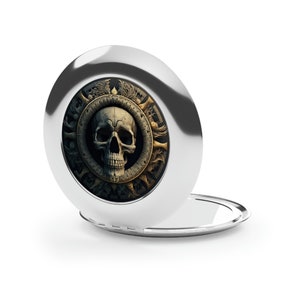 May include: A silver compact mirror with a skull design. The skull is surrounded by a circular design with gold accents. The mirror is open and the reflection is visible.
