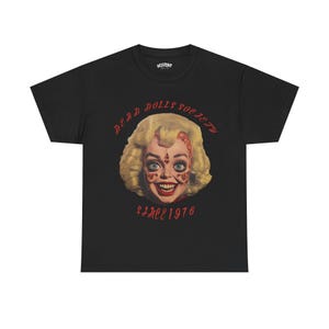 Può includere: Maglietta nera con una grafica rossa di un viso femminile con un sorriso inquietante. Il testo "Dead Dolls Society Since 1976" è stampato in rosso sulla maglietta.