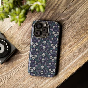 Puede incluir: Una funda de teléfono negra con un patrón repetido de calaveras blancas y flores moradas. La funda tiene un recorte para el objetivo de la cámara con tres agujeros.
