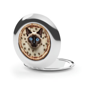 May include: A silver compact mirror with a cat's face peeking out from a pizza design on the front. The mirror is open and the reflective surface is visible.