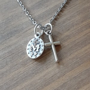 Puede incluir: Un collar plateado con una cadena, un peque&ntilde;o colgante de cruz y un colgante redondo con un emblema religioso. Los colgantes est&aacute;n conectados a la cadena por peque&ntilde;os bucles. El fondo es una superficie de madera.