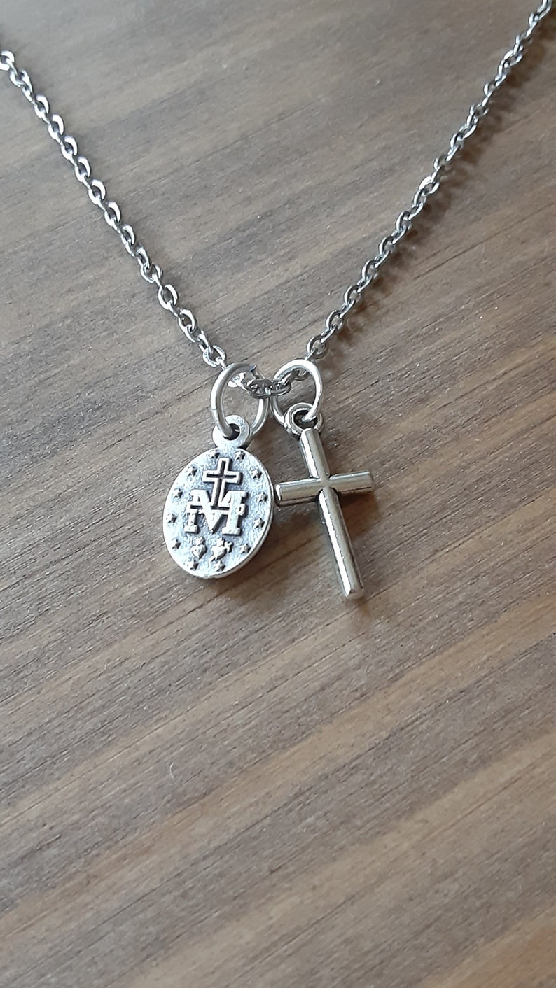 Puede incluir: Un collar plateado con una cadena, un peque&ntilde;o colgante de cruz y un colgante redondo con un emblema religioso. Los colgantes est&aacute;n conectados a la cadena por peque&ntilde;os bucles. El fondo es una superficie de madera.