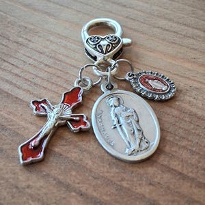 May include: A silver-toned metal charm featuring a heart-shaped lobster clasp attached to three pendants: a red enamel crucifix, an oval medal depicting Saint Dymphna with the text ST DYMPHNA, and a small oval medal with a red glitter inlay.