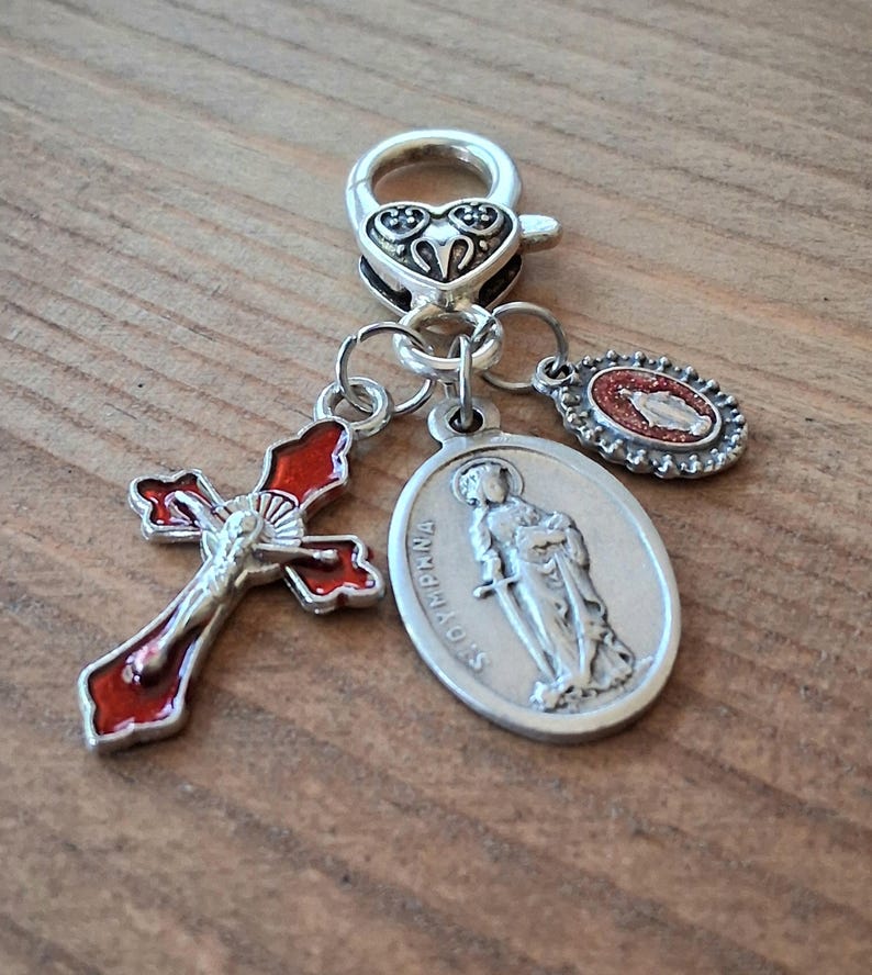 May include: A silver-toned metal charm featuring a heart-shaped lobster clasp attached to three pendants: a red enamel crucifix, an oval medal depicting Saint Dymphna with the text ST DYMPHNA, and a small oval medal with a red glitter inlay.