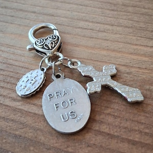 May include: A silver-toned metal charm featuring a heart-shaped lobster clasp, a small oval medal with an embossed letter M, a larger oval medal inscribed with the text PRAY FOR US ITALY, and a cross pendant with a textured, patterned surface.