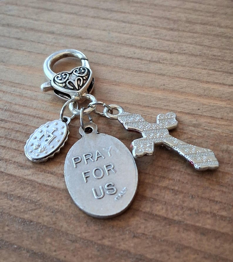 May include: A silver-toned metal charm featuring a heart-shaped lobster clasp, a small oval medal with an embossed letter M, a larger oval medal inscribed with the text PRAY FOR US ITALY, and a cross pendant with a textured, patterned surface.