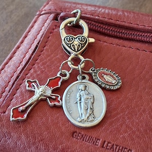 May include: A silver-toned metal lobster clasp charm featuring a heart-shaped top, attached to three dangling charms: a red and silver crucifix, a round silver medal depicting Saint Dymphna, and a small oval medal with a red center.
