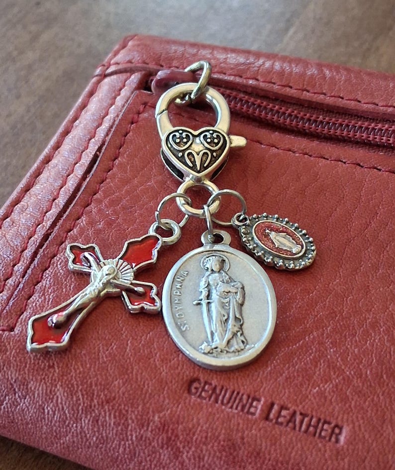 May include: A silver-toned metal lobster clasp charm featuring a heart-shaped top, attached to three dangling charms: a red and silver crucifix, a round silver medal depicting Saint Dymphna, and a small oval medal with a red center.