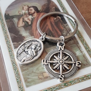 May include: A silver keychain with a compass charm and a charm featuring St. Christopher carrying a child. The charms are attached to a silver key ring. The keychain is displayed on a card with a picture of St. Christopher.