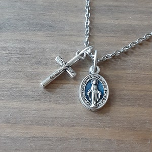 Puede incluir: Collar plateado con un colgante de cruz y un colgante circular con una imagen religiosa azul y blanca. La cadena es delicada y los colgantes est&aacute;n detallados. El collar descansa sobre una superficie de madera.