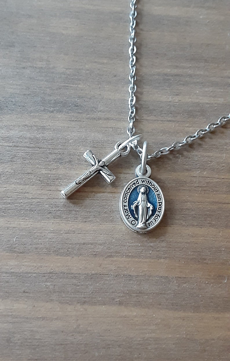 Puede incluir: Collar plateado con un colgante de cruz y un colgante circular con una imagen religiosa azul y blanca. La cadena es delicada y los colgantes est&aacute;n detallados. El collar descansa sobre una superficie de madera.