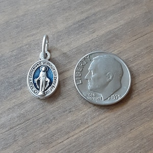 Puede incluir: Un peque&ntilde;o colgante ovalado plateado con un fondo de esmalte azul y una representaci&oacute;n de la Virgen Mar&iacute;a. El colgante est&aacute; junto a una moneda de diez centavos estadounidense para comparar el tama&ntilde;o. El colgante tiene la inscripci&oacute;n "Oh Mar&iacute;a, concebida sin pecado, ruega por nosotros."