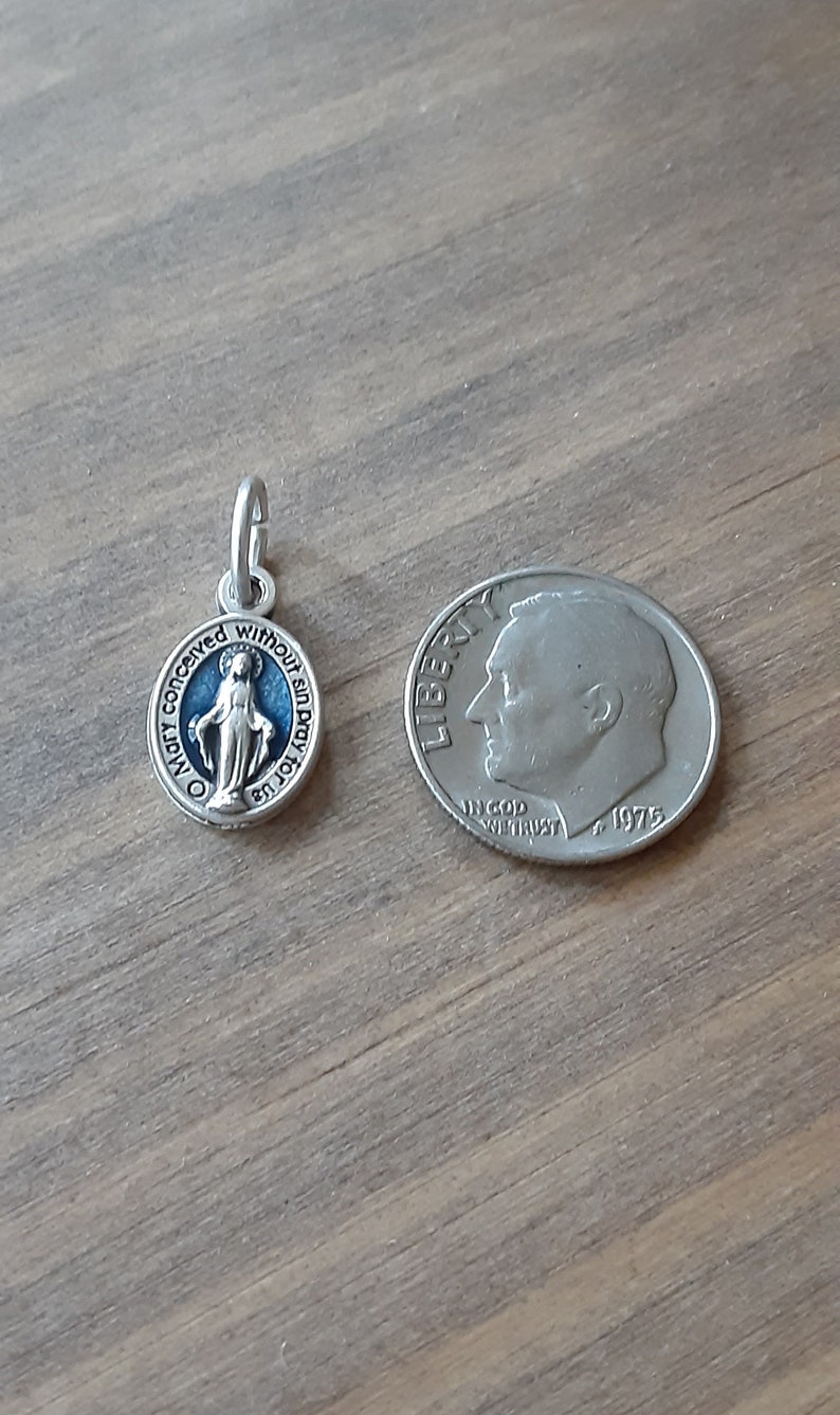 Puede incluir: Un peque&ntilde;o colgante ovalado plateado con un fondo de esmalte azul y una representaci&oacute;n de la Virgen Mar&iacute;a. El colgante est&aacute; junto a una moneda de diez centavos estadounidense para comparar el tama&ntilde;o. El colgante tiene la inscripci&oacute;n "Oh Mar&iacute;a, concebida sin pecado, ruega por nosotros."