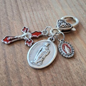 May include: A silver-toned metal charm featuring a heart-shaped lobster clasp attached to three pendants: a red enamel crucifix, an oval medal embossed with the figure of Saint Dymphna, and a smaller oval medal with a red glitter inlay of a figure.